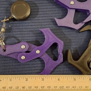 May include: Assortment of 3D-printed keychains in orange, purple, and black. Each keychain features a unique design, including a claw-like shape and a retractable reel. A wooden ruler with inch markings is in the foreground.