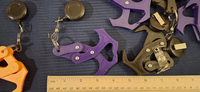 May include: Assortment of 3D-printed keychains in orange, purple, and black. Each keychain features a unique design, including a claw-like shape and a retractable reel. A wooden ruler with inch markings is in the foreground.