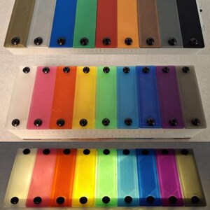 May include: A set of three colorful xylophones. Each xylophone has a white base and a series of rectangular bars in various colors, secured with black screws. The bottom xylophone has the text "Radiant Artifacts" on the base.