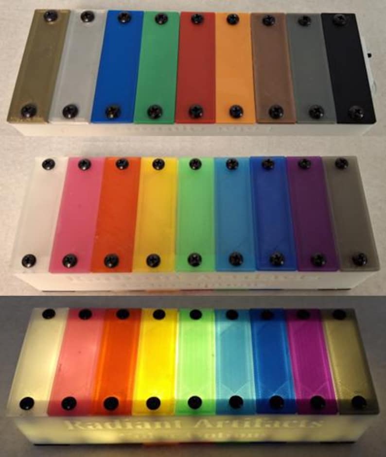 May include: A set of three colorful xylophones. Each xylophone has a white base and a series of rectangular bars in various colors, secured with black screws. The bottom xylophone has the text "Radiant Artifacts" on the base.