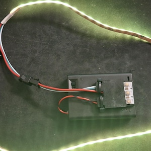 May include: A flexible LED light strip emitting a warm white glow, connected by thin red, white, and blue wires to a rectangular black battery pack. The battery pack features a small sliding power switch and exposed internal wiring components.