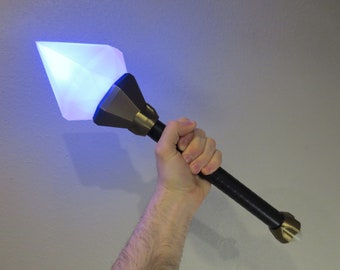May include: A blue glowing diamond-shaped object attached to a black handle with a gold metal base. The handle is held by a hand.
