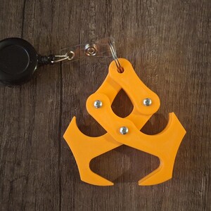 May include: An orange, articulated keychain accessory with a retractable black reel. The accessory has a claw-like design with multiple pivoting joints and silver rivets. The item is shown on a dark wood grain surface.