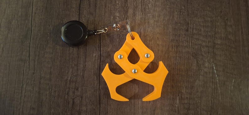 May include: An orange, articulated keychain accessory with a retractable black reel. The accessory has a claw-like design with multiple pivoting joints and silver rivets. The item is shown on a dark wood grain surface.