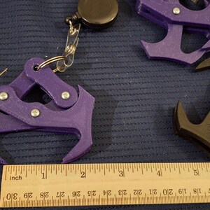 May include: Assortment of 3D-printed grappling hooks in orange, purple, and black, each attached to a retractable reel. A wooden ruler with inch markings is placed near the hooks. The hooks are approximately 3 inches long.