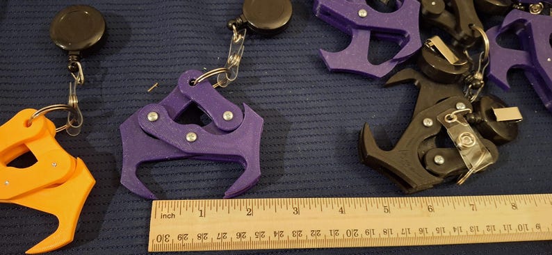 May include: Assortment of 3D-printed grappling hooks in orange, purple, and black, each attached to a retractable reel. A wooden ruler with inch markings is placed near the hooks. The hooks are approximately 3 inches long.