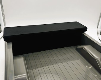 May include: A gray truck bed featuring a ribbed floor and two wheel wells, topped with a solid black rectangular storage box mounted across the front panel. The interior surface is smooth and glossy, showing two circular holes on the floor.