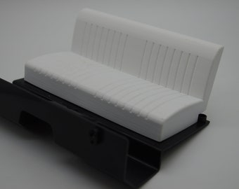 May include: White plastic model car seat with a black plastic base. The seat has a ribbed texture and is designed for a model car.