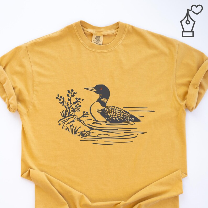 May include: Mustard yellow t-shirt featuring a black and white illustration of a loon swimming in water. The loon has a black head, a white neck, and a patterned body. The design includes a small bush on the left side.
