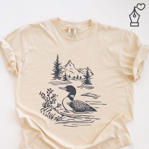 May include: Cream-colored t-shirt featuring a black and white illustration of a loon swimming in a lake with mountains and trees in the background. The shirt has a classic crew neck and short sleeves.