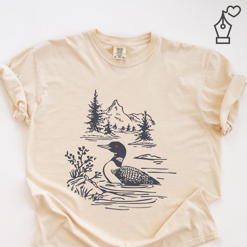 May include: Cream-colored t-shirt featuring a black and white illustration of a loon swimming in a lake with mountains and trees in the background. The shirt has a classic crew neck and short sleeves.