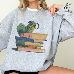 Peut inclure: Un sweat-shirt gris clair avec une illustration colorée d'une chenille verte portant des lunettes, perchée sur une pile de livres. Les livres sont dans des tons de bleu, de rose et de jaune. Le sweat-shirt a un col rond.