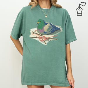 May include: A teal green t-shirt featuring a graphic of a pigeon on a nest. The pigeon has green, blue, and purple feathers. The nest is made of twigs and red string. The shirt has a relaxed fit and short sleeves.