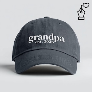 May include: A dark gray baseball cap with the word "grandpa" embroidered in white, along with "est. 2026". The cap has a curved brim and a classic six-panel design. A black and white illustration of a pen with a heart is in the upper right corner.