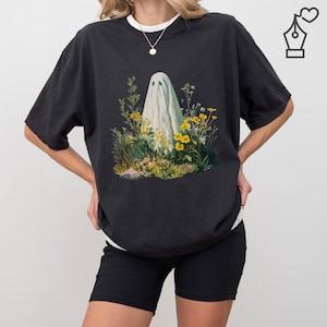 Puede incluir: Camiseta negra con una ilustración de un fantasma entre flores silvestres. El fantasma está envuelto en una sábana blanca, rodeado de follaje amarillo y verde. La camiseta tiene un corte holgado y cuello redondo blanco.