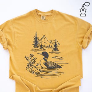 Puede incluir: Camiseta de color amarillo mostaza con una ilustración en blanco y negro de un somorgujo nadando en el agua, con un paisaje de montañas y pinos al fondo. La camiseta tiene cuello redondo y mangas cortas.