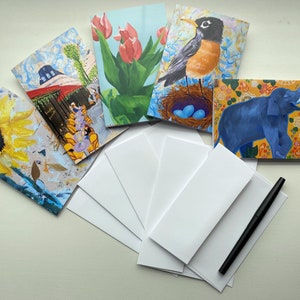 May include: A set of blank greeting cards with envelopes and a black pen. The cards feature colorful illustrations of a sunflower, a robin in a nest, tulips, a blue elephant, and a Parisian cafe scene.