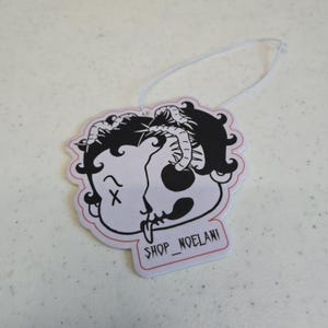 May include: A white air freshener featuring a black and white illustration of a skull-like face with a snake and horns. It has a red border and a white string. The text "SHOP_NOELANI" is printed at the bottom of the air freshener.