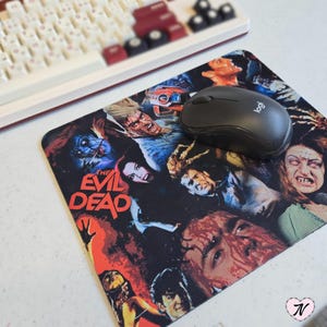 Op de afbeelding: Een zwarte muismat met een afbeelding van personages uit de film "The Evil Dead". De muismat bevat een zwarte computermuis met het woord "Logi" erop.