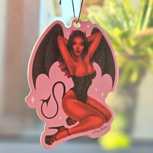 Puede incluir: Ambientador de coche rosa con una mujer diablo roja con alas negras y cola. La mujer lleva un atuendo negro y rojo y tiene una corona de cuernos. El ambientador tiene detalles de purpurina y el texto "@Shop_Moolau" en la parte inferior.