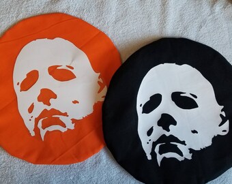 Könnte beinhalten: Zwei runde Stoffkissen mit einer weißen Silhouette der Maske von Michael Myers aus der Halloween-Reihe. Ein Kissen ist orange und das andere ist schwarz.
