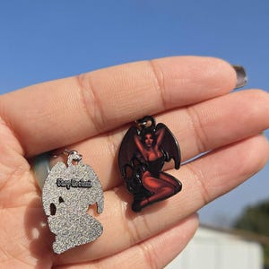 May include: Two metal pendants. One is silver with the text "Shop Useless" and a silhouette design. The other is black with a red devil woman design. Both pendants have a small loop for attaching to a chain or other item.
