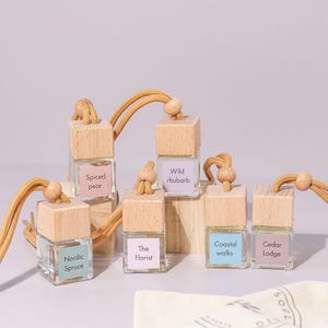 May include: Six small glass bottles with wooden caps and string hangers. Each bottle has a different scent label: Spiced Pear, Wild Rhubarb, The Florist, Nordic Spruce, Coastal Walks, and Cedar Lodge.