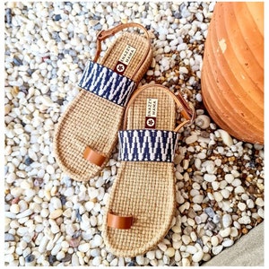 May include: A pair of brown woven sandals with blue and white patterned straps. The sandals have a leather toe strap and a leather ankle strap with the text "Snazzy Mama" printed on it.