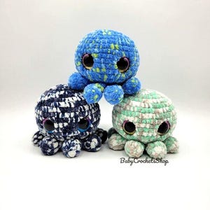 May include: Three handmade crochet octopus toys in blue, navy, and green. Each octopus has large, round, black eyes and eight tentacles. The blue octopus is on top, the navy one in the middle, and the green one at the bottom. The text "BabyCrochetsShop" is visible.