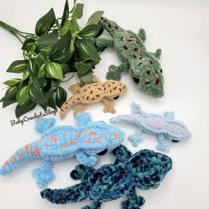 May include: A collection of handmade crochet gecko plushies in various colors, including blue, green, and tan. The plushies have detailed features and are arranged with green foliage. The text "Baby Crochets Shop" is visible.