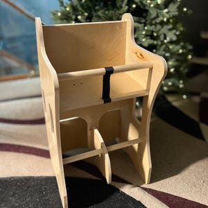May include: A natural wood-toned, wooden learning tower with a rectangular frame. It features a shelf, a footrest, and a black strap. The tower is designed to help children reach countertops and other elevated surfaces.