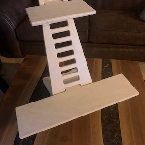 May include: A light-colored wooden standing desk converter with a rectangular base and a top platform. The desk has a series of rectangular cutouts along its central support structure. The desk is designed to sit on top of a table.