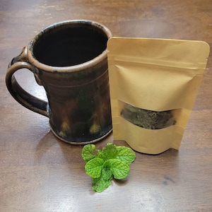 May include: A brown ceramic mug with a handle, a brown paper bag with a clear window showing dried herbs inside, and a sprig of fresh green mint leaves on a wooden surface.