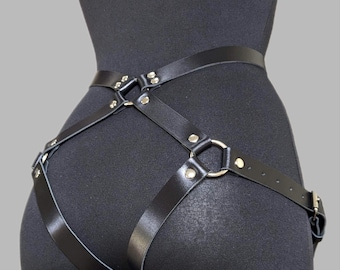 May include: A black leather body harness featuring adjustable straps, silver-toned metal rings, and rivets. The harness consists of a waist belt connected to leg loops with intersecting straps, secured by metal buckles and multiple adjustment holes.