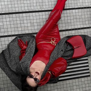 Könnte beinhalten: Ein glänzendes rotes Latexkleid mit einer goldenen Taillenkette, roten Latexhandschuhen, roten spitzen Schuhen und einer roten Handtasche. Ein grauer Tweedmantel liegt über dem Outfit. Das Ensemble umfasst eine dunkle Sonnenbrille.