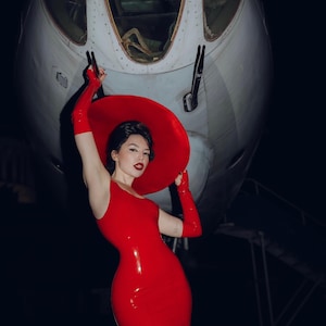Könnte beinhalten: Ein ärmelloses, eng anliegendes rotes Midikleid aus Latex, kombiniert mit einem roten Hut mit breiter Krempe und passenden ellbogenlangen roten Handschuhen. Das Ensemble zeichnet sich durch eine glänzende Oberfläche und kräftiges Rot aus.