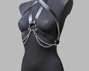 May include: A black faux leather body harness featuring adjustable straps that cross at the chest and neck. The harness is accented with silver-toned metal hardware, including circular rings and two draped silver chains hanging from the front.