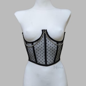 May include: A black corset top featuring sheer mesh fabric with a black polka dot pattern. The garment includes black structural boning, a silver-toned metal chain draped across the front, and the word Nova printed in small gray letters.