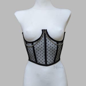 May include: A black corset top made of sheer mesh fabric with a black polka dot pattern. The garment features vertical black boning, a sweetheart neckline, and a silver-toned metal chain draped across the front. The word Nova is printed in small grey letters near the bottom right.