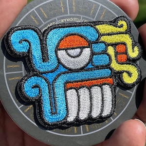 Pode incluir: Um remendo bordado azul, amarelo, laranja e branco que representa um rosto estilizado com um olho grande. O remendo está sobre um fundo preto com um círculo dourado e um padrão de linhas douradas.