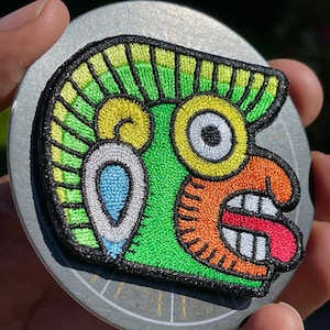 Puede incluir: Parche bordado con un rostro estilizado de colores con una boca grande abierta y una lengua roja. El rostro tiene un fondo verde con detalles amarillos y blancos. El parche es circular y tiene un borde negro.