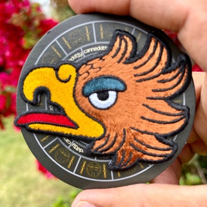 May include: A brown and yellow embroidered patch of a bird's head with a red beak and a blue eye. The patch is on a black circular background with gold lettering.