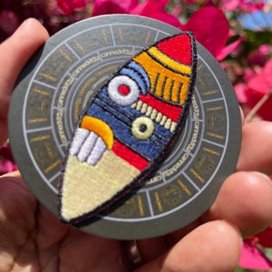 May include: Embroidered patch with a colorful abstract design. The patch features red, yellow, blue, and white stitching. The patch is on a gray circular surface with the text "cannedkitty" repeated around the edge.