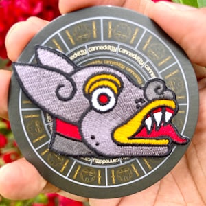 May include: A gray embroidered patch of a snarling wolf-like creature with red, yellow, and white details. The patch is on a black circular background with gold accents.