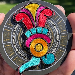 May include: A colorful embroidered patch with a yellow, red, pink, and blue design. The patch is circular and has a black outline. The patch is on a black background with the text "cannedkitty" repeated around the edge.