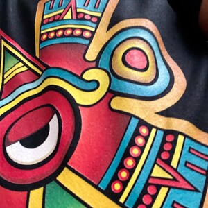 以下が含まれることがあります： 大きな目がついた赤、黄、青の顔を描いた、カラフルなアステカ風のデザインが施された黒いTシャツです。