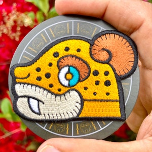 May include: A yellow embroidered patch with a cartoon animal with black spots, a white and blue eye, and an orange ear. The patch is on a black background with a gold circle design.