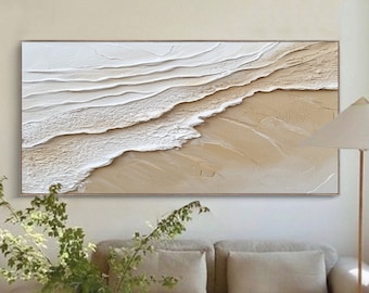 May include: Abstract painting of white waves crashing on a beige sandy beach. The painting is framed and hanging on a wall above a beige sofa.