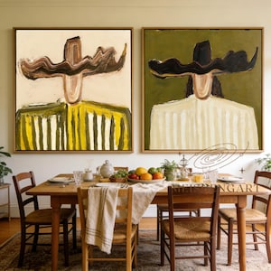 May include: Two framed abstract paintings. One features a figure with a brown head and yellow and green striped shirt on a cream background. The other has a black head and white striped shirt on a green background. Both are displayed above a wooden dining table.