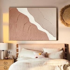 May include: Abstract painting with a textured, layered design in shades of pink and white. The painting is framed in a gold frame and hangs above a bed with white bedding.